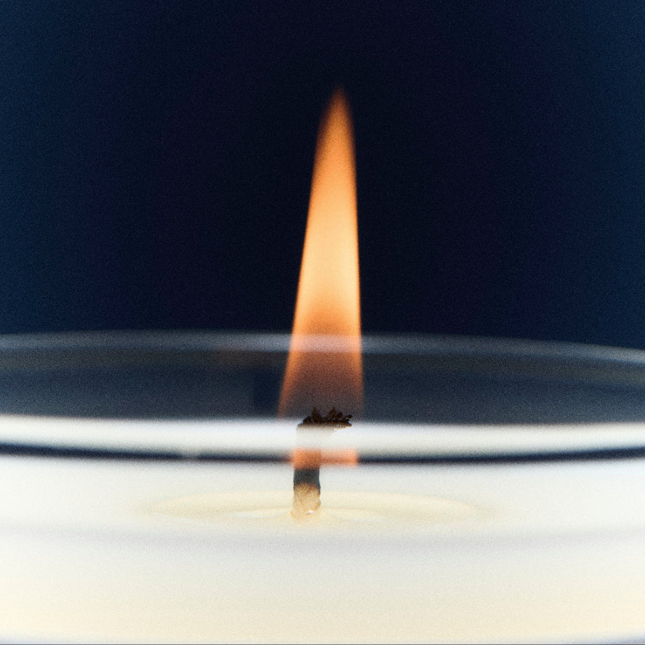 A close-up shot of a lit candle with a single wick and a tall orange flame against a dark navy blue background.