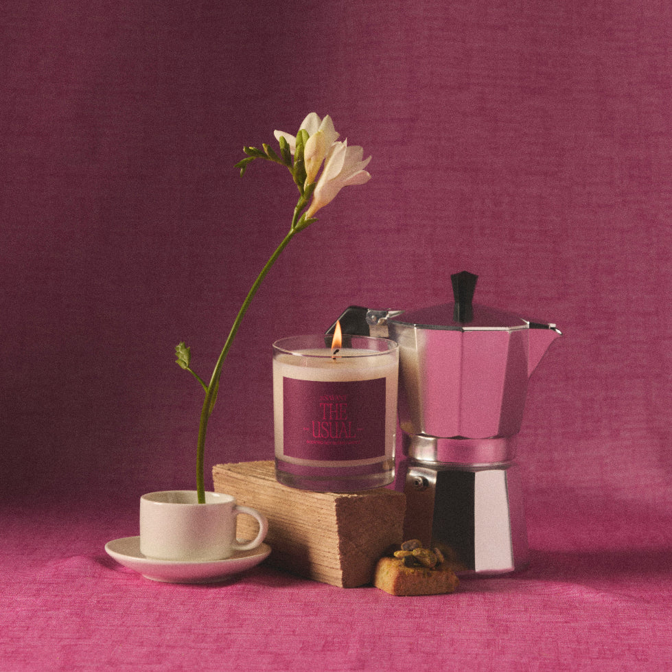 A lit glass jar candle with a dark purple label sits on a wooden block beside a silver moka pot, a small white cup and saucer with a single white flower, and a small pastry, all set against a textured purple backdrop.